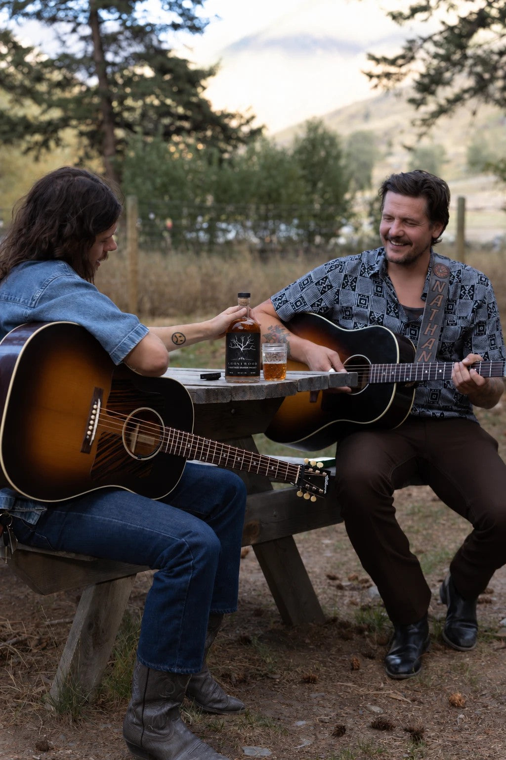 Two friends with guitars at a picnic table, a bottle of Ghostwood 3 Brothers Bourbon between them