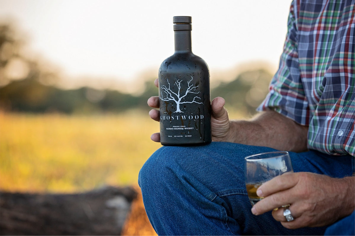 Ghostwood Original Blended Bourbon held on a knee with a pour, Montana field at sunset